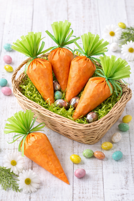 DIY Easter carrot treat bags made with orange tissue paper and candy eggs displayed in a wicker Easter basket